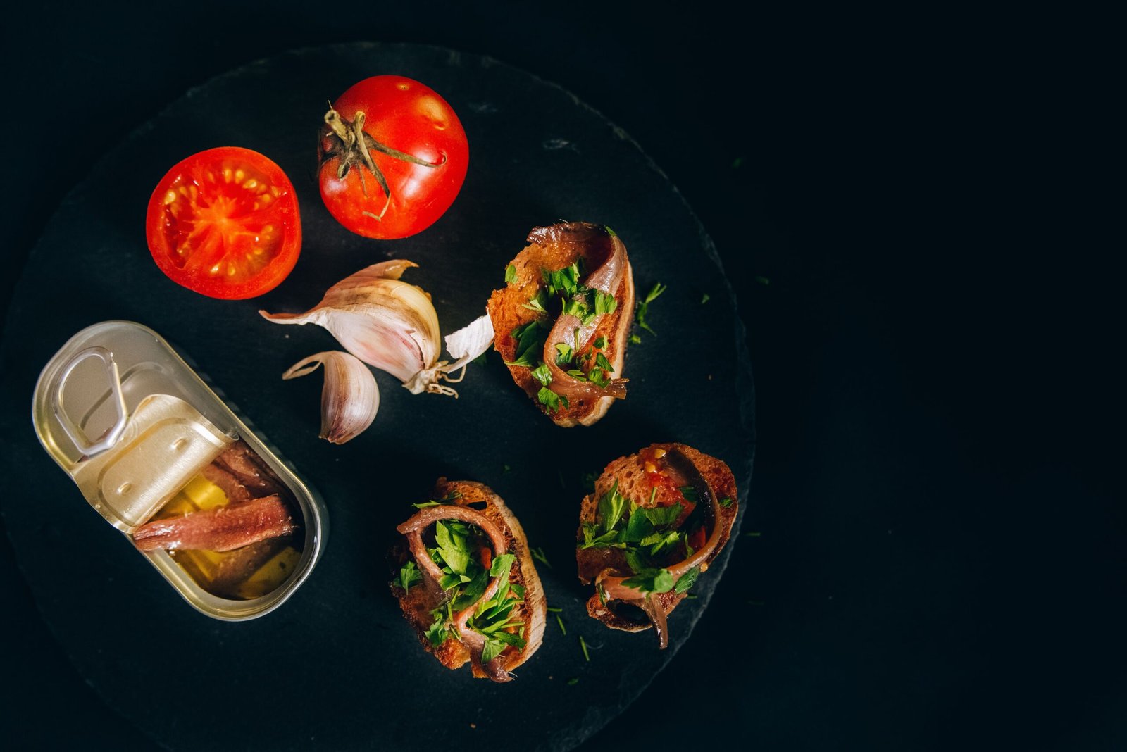Anchovies served on toasted bread with tomatoes, garlic, and fresh parsley beside an open tin.